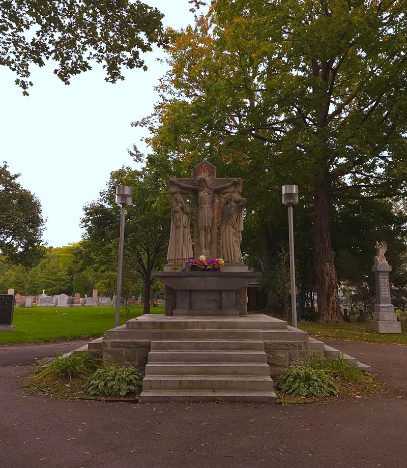 Calvaire de pierre : le Christ en croix entre deux statues, sur un socle à plusieurs marches, au milieu d'un rond-point bordé de grands arbres.