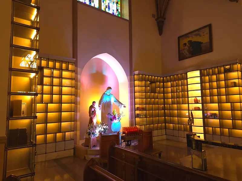 Niches vitrées baignées de lumière dorée autour d'une statue du Christ aux bras ouverts, sous un vitrail, avec les bancs au premier plan.