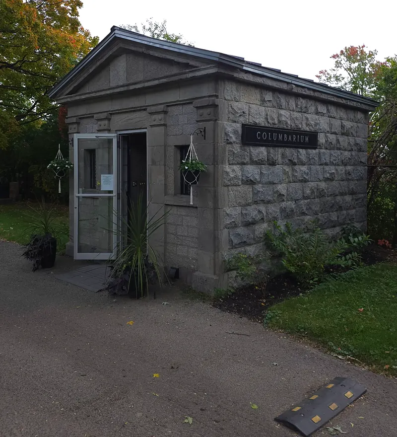 Petit bâtiment de pierre grise à fronton, portant la plaque « Columbarium », sa porte vitrée ouverte sur l'allée.