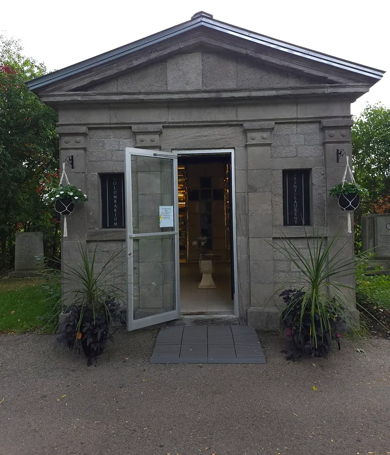 Façade de pierre grise du columbarium extérieur, porte vitrée ouverte, plantes en pot et paniers suspendus de chaque côté.