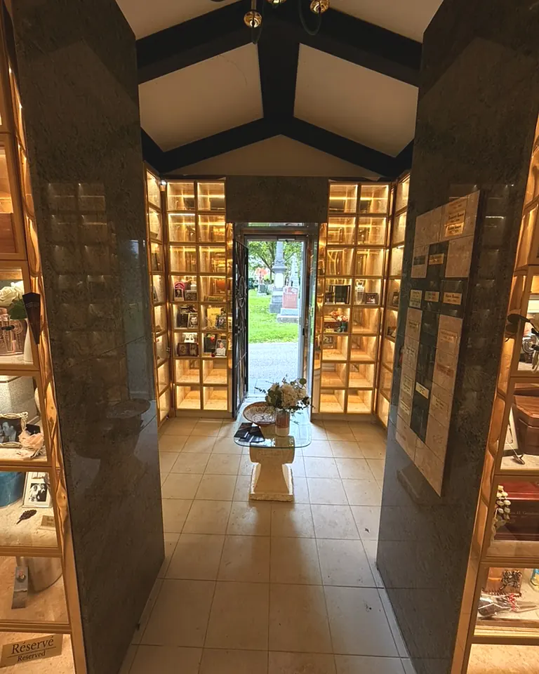 Intérieur du columbarium : niches vitrées éclairées de chaque côté, colonnes de granit et porte ouverte sur le cimetière.